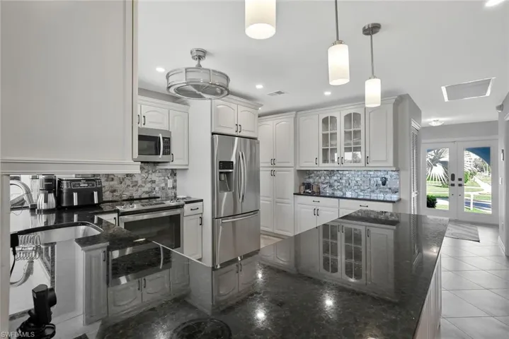 Kitchen with dark stone countertops, stainless steel appliances, glass insert cabinets, backsplash, and white cabinets