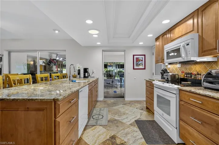 Kitchen with white appliances, decorative backsplash, brown cabinets, recessed lighting, and light stone countertops
