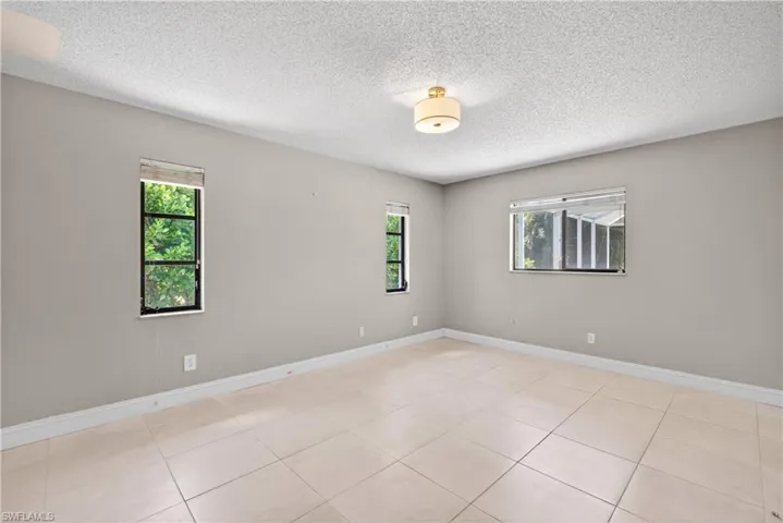 Spacious room featuring neutral painted walls, tile flooring, three windows with natural light, and a flush mount ceiling light fixture