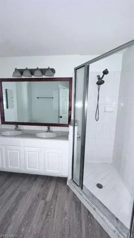 Bathroom featuring a glass-enclosed shower, dual vanity with white cabinetry, large framed mirror, and wood-finish flooring