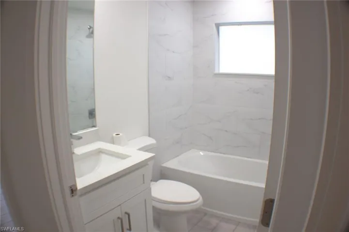 Bathroom featuring a white vanity with integrated sink, white tiled flooring, a standard toilet, and a bathtub with white subway tile surround and a window