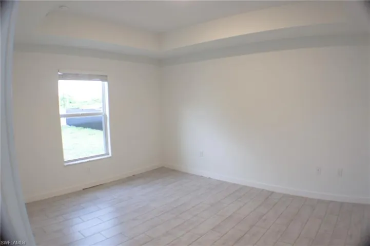 Spacious room featuring light-toned wood-finish flooring, a tray ceiling with recessed lighting, and a window with blinds