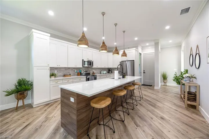 Modern kitchen featuring a large island with a white countertop and wood-finish paneling, pendant lighting, white cabinetry, stainless steel appliances, and wood-finish flooring