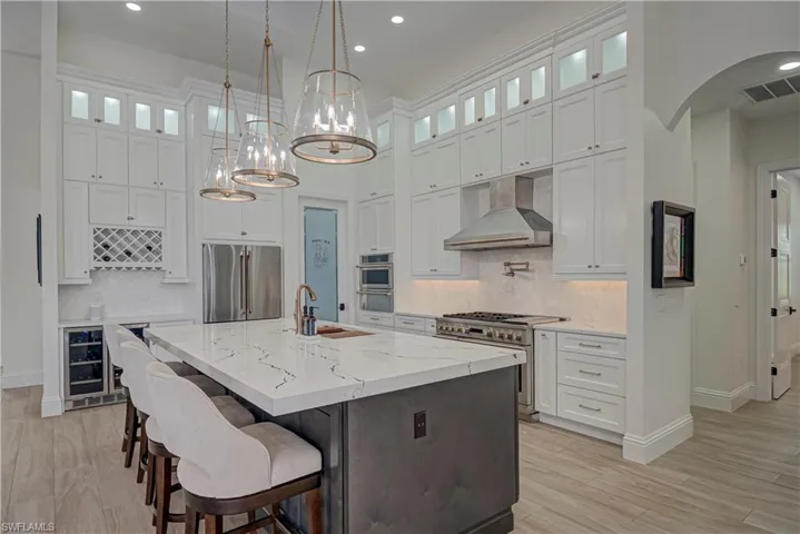 Kitchen featuring glass insert cabinets, white cabinetry, decorative light fixtures, wall chimney exhaust hood, and a towering ceiling