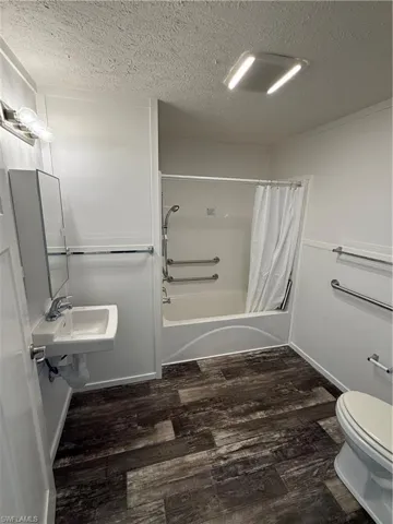 Wood-finish flooring throughout the bathroom featuring a wall-mounted sink, a built-in tub with a shower fixture, and multiple grab bars