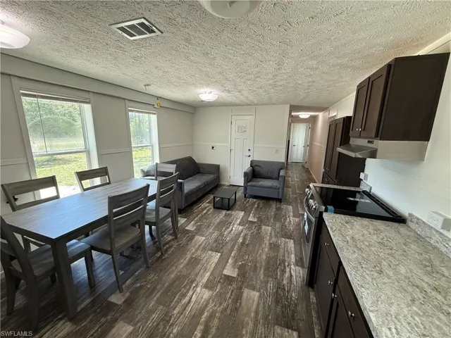 Open-concept living space featuring wood-finish flooring throughout, a full kitchen with dark cabinetry and granite-style countertops, dining area with seating for multiple guests, and a comfortable lounge zone