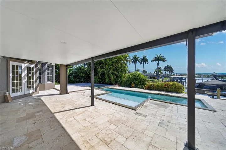 Expansive covered patio with tile flooring, featuring a swimming pool, and views of palm trees and the distant water