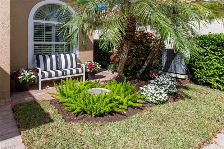 Stucco exterior featuring an arched window with decorative molding, paver patio, and professional landscaping with mature palm trees and flowering shrubs