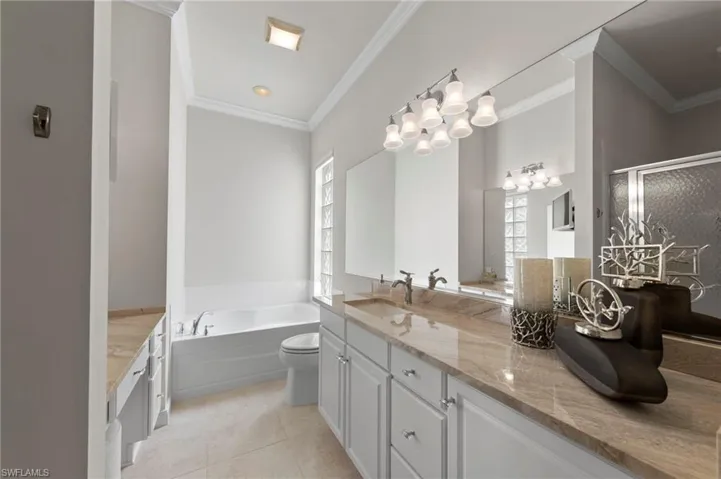 Dual vanity with stone-finish countertops, glass block window, soaking tub, crown molding, and polished metallic light fixtures