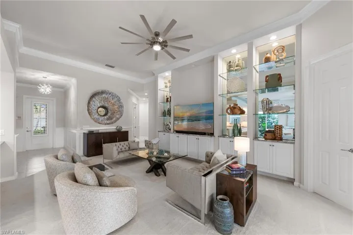 Living area featuring crown molding, built-in cabinetry with glass shelving, mirror backsplashes, and a multi-blade ceiling fan