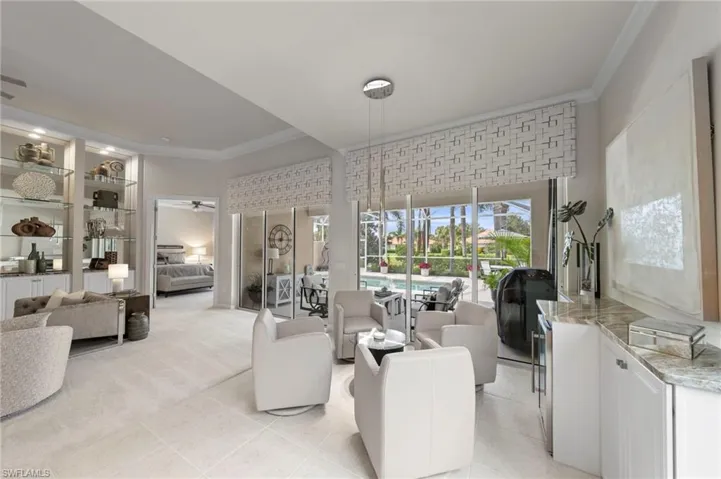 Living space featuring neutral stone-finish tile flooring, built-in floor-to-ceiling glass display shelving, a dry bar with stone-surfaced countertop, and floor-to-ceiling sliding glass doors opening to a screened pool enclosure
