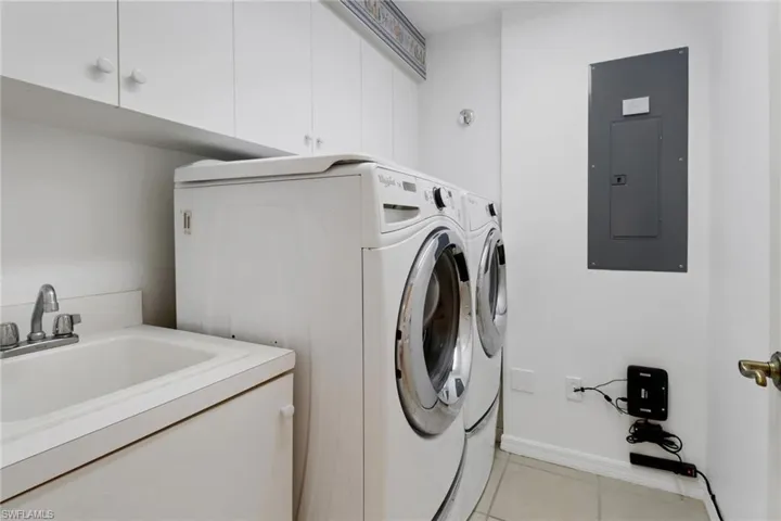 Clothes washing area with electric panel, washer and dryer, sink, light tile patterned flooring, and cabinets