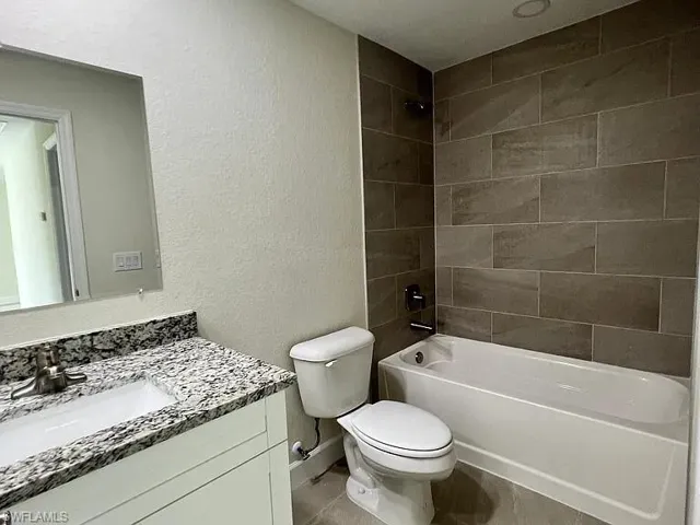 Bathroom featuring a vanity with stone-patterned countertop, rectangular undermount sink, wall-mounted mirror, white cabinetry, and a bathtub with large-format tile surround