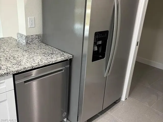 Stainless steel side-by-side refrigerator, matching stainless steel dishwasher, speckled stone-finish countertops, and large-format tile flooring
