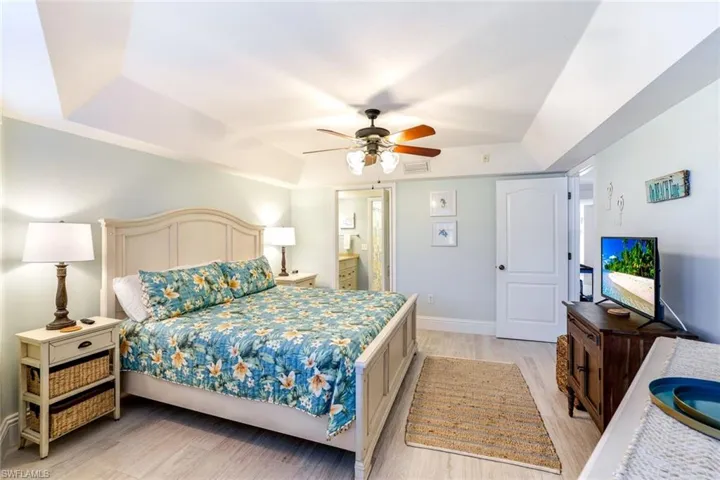 Bedroom with a raised ceiling, light wood finished floors, connected bathroom, and a ceiling fan