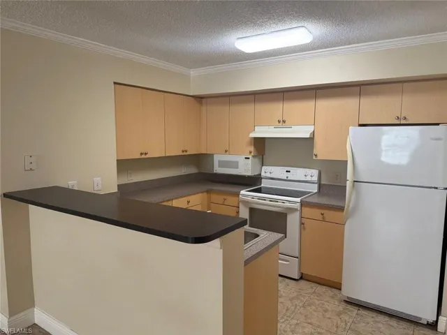 Kitchen with white appliances, a peninsula, dark countertops, a textured ceiling, and light wood finish cabinetry