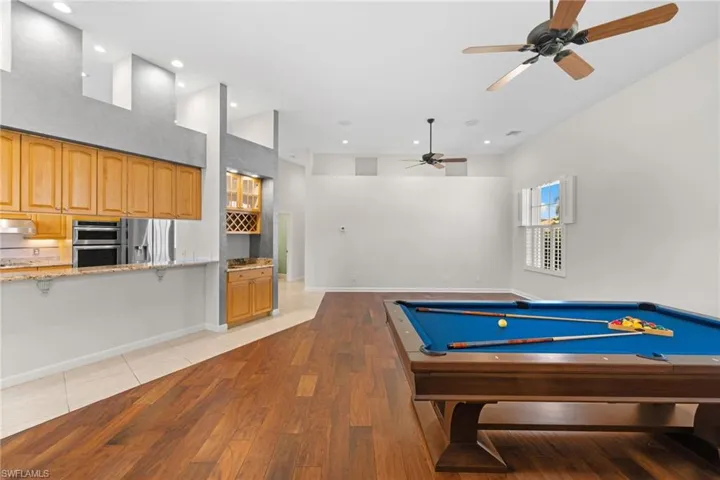 Open-concept living space featuring wood-finish flooring, a kitchen pass-through with a granite-look counter, built-in double ovens, a wine rack, and high ceilings with recessed lighting and ceiling fans