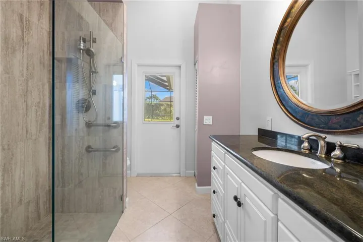 Bathroom #2 with access to outdoor living space featuring a frameless glass shower enclosure, wood-finish shower wall tile, brushed nickel fixtures, a single vanity with dark stone countertop, and a large decorative mirror