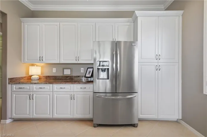 Kitchen with white cabinetry, stainless steel fridge, crown molding, light tile patterned floors, and dark stone counters