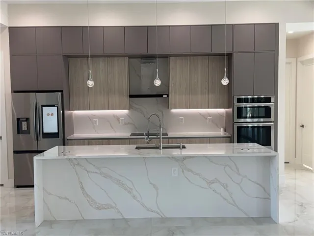 Kitchen with an island with sink, stainless steel appliances, sink, light stone counters, and wall chimney range hood