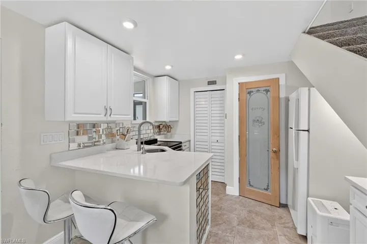 White shaker-style cabinetry, mosaic tile backsplash, stainless steel faucet, light-toned countertop, and brown tile flooring