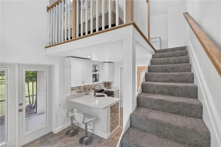 Open-concept layout with a white railing balcony and wood-finish handrail, bright white cabinetry, a kitchen peninsula with seating, and a large glass-paneled door