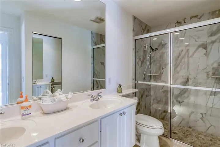 Bathroom featuring a dual vanity with undermount sinks and chrome fixtures, a spacious walk-in shower with glass enclosure and stone-finish tile surround, and a toilet with a convenient shelf