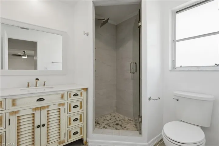 Bathroom featuring a distressed wood-finish vanity with a white countertop and undermount sink, a frameless glass shower enclosure with a rain shower head and pebble-tiled floor, and a white toilet
