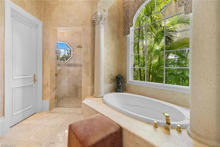 Arched window overlooking tropical foliage, built-in soaking tub with brass fixtures, separate glass-enclosed shower with a bench, ornate architectural column, and light-toned tile flooring
