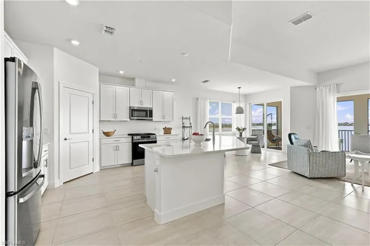 Open-concept living space with light-toned tiled flooring, a kitchen island with a white countertop, white cabinetry with stainless steel appliances, and glass sliding doors leading to an outdoor balcony