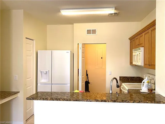 Kitchen with white appliances, a peninsula, brown cabinetry, dark stone countertops, and electric panel