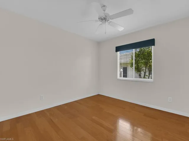 Room featuring light-toned walls, wood-finish flooring, a ceiling fan, and a window with horizontal blinds and a dark valance