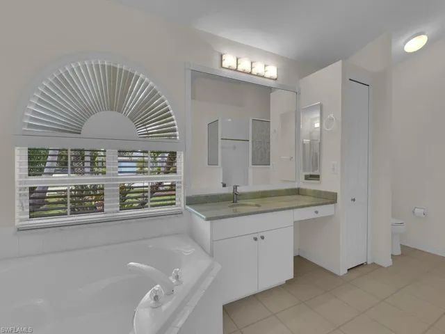 Soaking tub with white fixtures, an arched window featuring horizontal blinds, and a light green countertop vanity