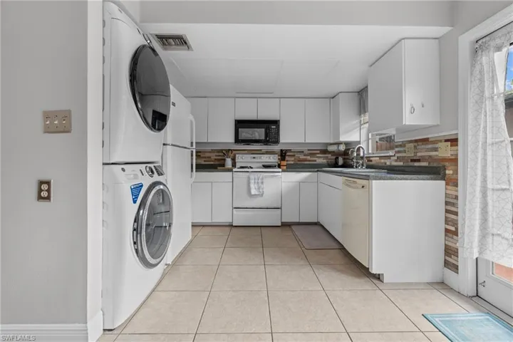 Kitchen featuring white appliances, light tile patterned floors, dark countertops, stacked washer and clothes dryer, and white cabinetry