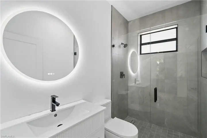 Bathroom featuring a back-lit round mirror, a white vanity with an integrated sink and matte black faucet, a walk-in shower with glass enclosure, and grey tile walls and flooring