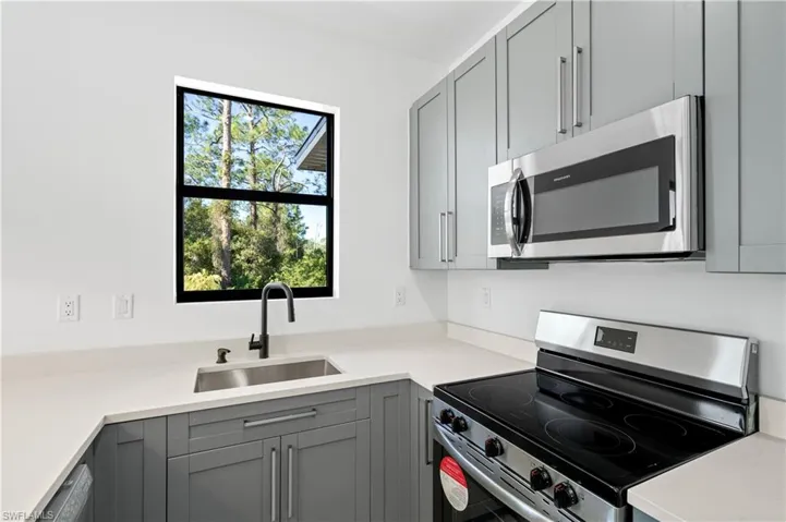 Modern kitchen featuring gray cabinetry, stainless steel appliances, light-toned countertops, a black-framed window, and a matte black faucet