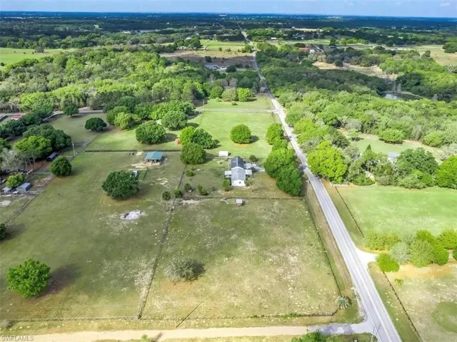 Expansive property featuring a single-story residence, multiple outbuildings, mature trees, and open fields, all bordered by a paved road