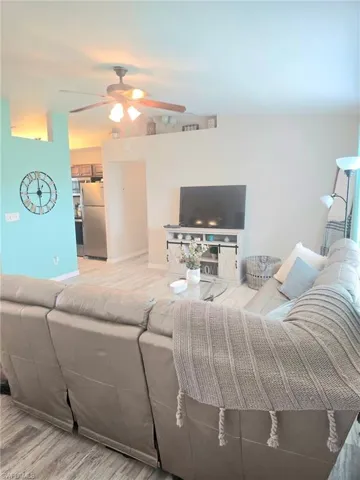 Living room with a ceiling fan, light wood finished floors, and baseboards including TV stand and television and wall hangings
