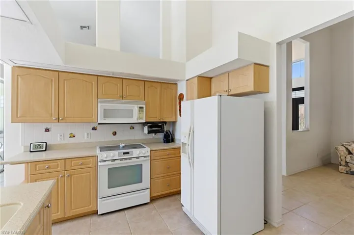 Kitchen featuring light wood finish cabinets, white appliances, light tile patterned floors, decorative backsplash, and a high ceiling