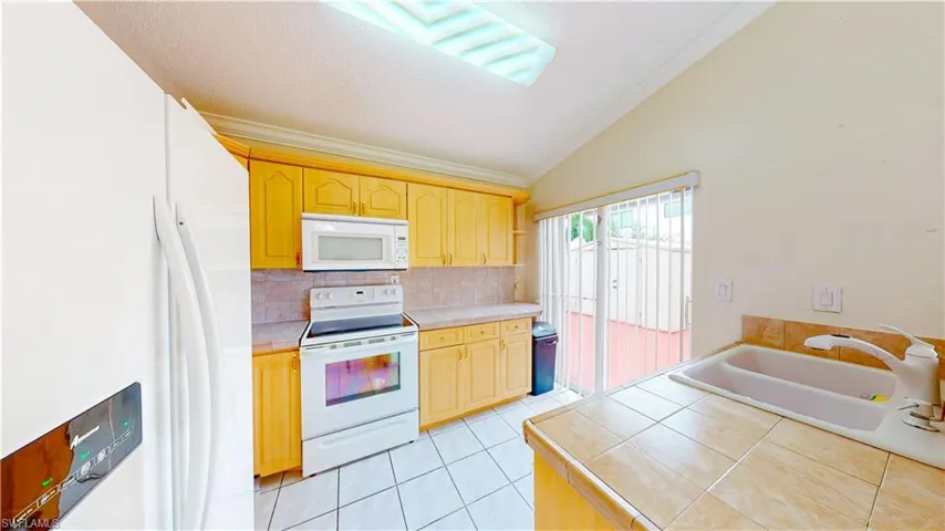 Kitchen featuring tile countertops, white appliances, vaulted ceiling, decorative backsplash, and light brown cabinets