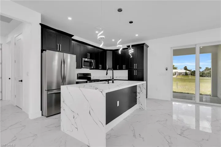 Kitchen with dark cabinets, light stone countertops, stainless steel appliances, a center island with sink, and decorative light fixtures