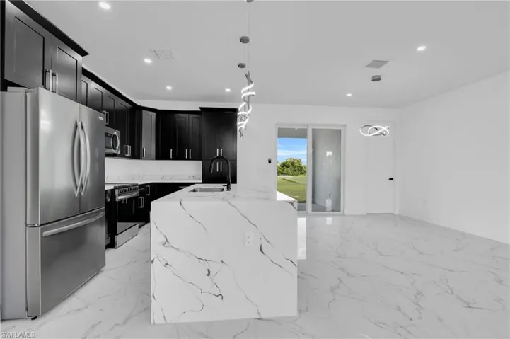 Kitchen featuring stainless steel appliances, dark cabinets, light stone countertops, an island with sink, and decorative light fixtures