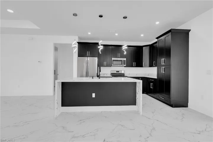 Kitchen featuring light stone counters, dark cabinetry, stainless steel appliances, a kitchen island with sink, and decorative light fixtures