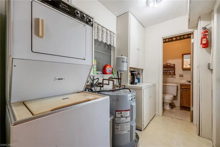 Clothes washing area featuring electric water heater, a textured ceiling, light floors, stacked washer / drying machine, and laundry area