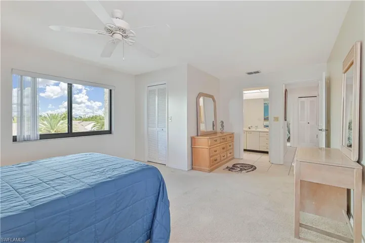 Carpeted bedroom featuring a ceiling fan, a large window with exterior foliage, and an adjacent bathroom with tile flooring and a vanity