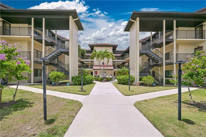 Multi-story residential complex featuring private balconies, exterior staircases, manicured grounds, mature landscaping, and concrete pathways