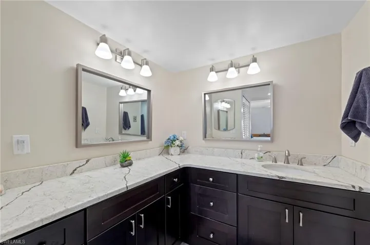 L-shaped vanity featuring dark wood cabinetry, veined stone-finish countertop, dual under-mount sinks, framed wall mirrors, and multi-bulb vanity lighting