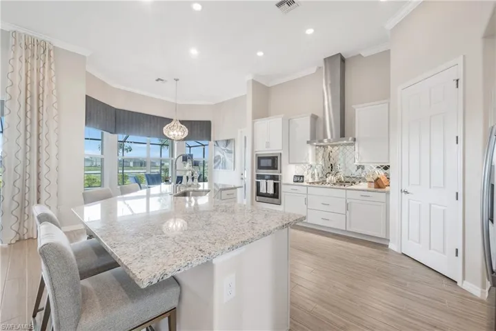 Kitchen with appliances with stainless steel finishes, light flooring, a kitchen island with sink, wall chimney range hood, and white cabinets