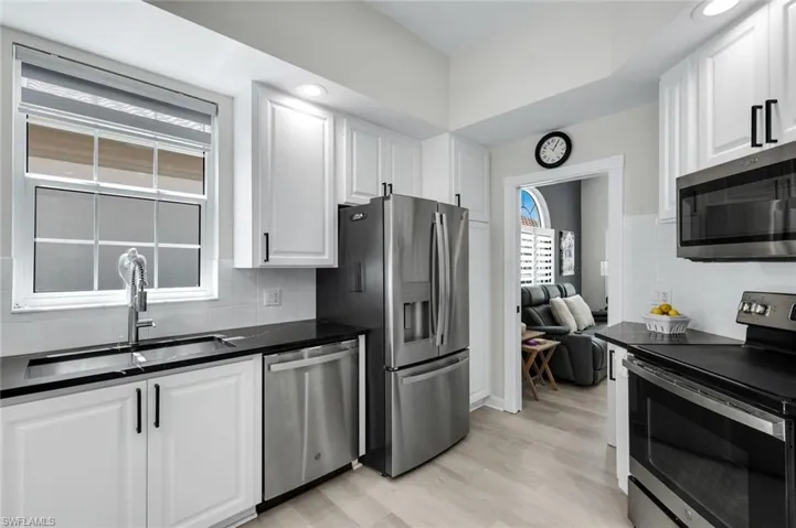 White cabinetry with black hardware, stainless steel appliances, dark countertops, white tile backsplash, and light wood-finish flooring