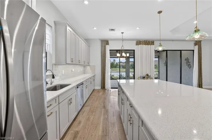 Spacious kitchen featuring a large central island with a light-toned countertop, stainless steel appliances, extensive white cabinetry, wood-finish flooring, and pendant lighting